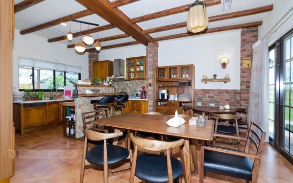 Una cocina amplia con vigas de madera, un comedor con una mesa grande y sillas, y una pared de ladrillo con estanterías.