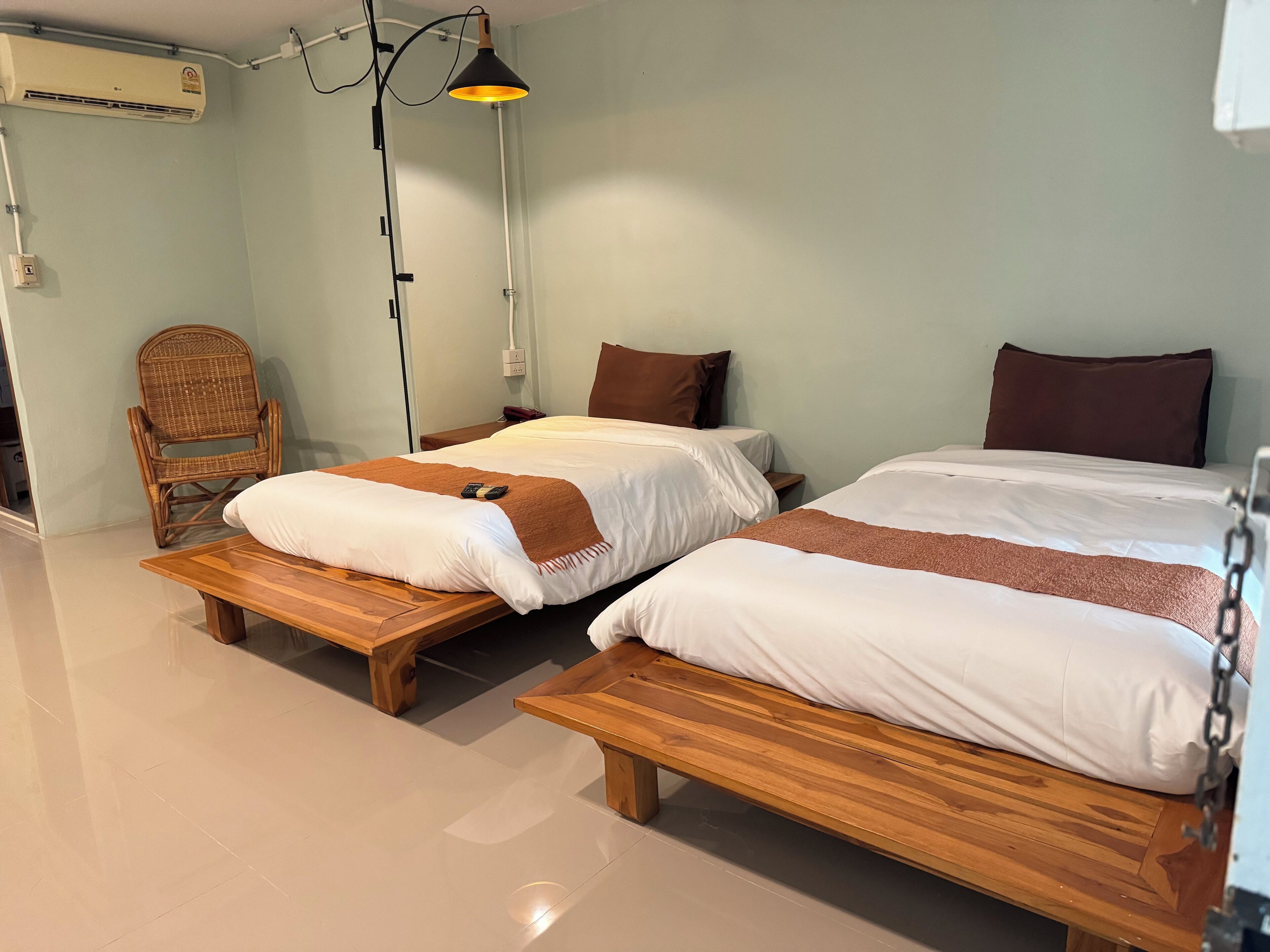 Habitación de hotel con dos camas, un banco de madera, una silla de mimbre y una lámpara de pie.