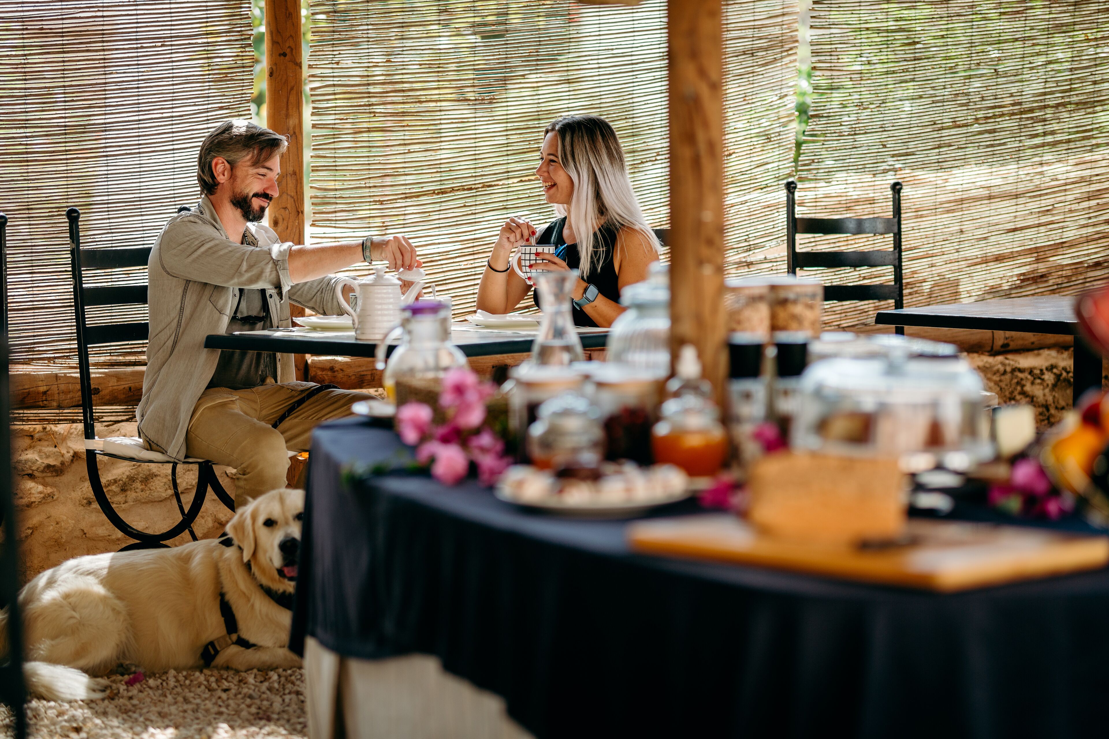 Un comedor con una mesa dispuesta para dos personas, rodeada de diversos alimentos y un perro acostado en el suelo.