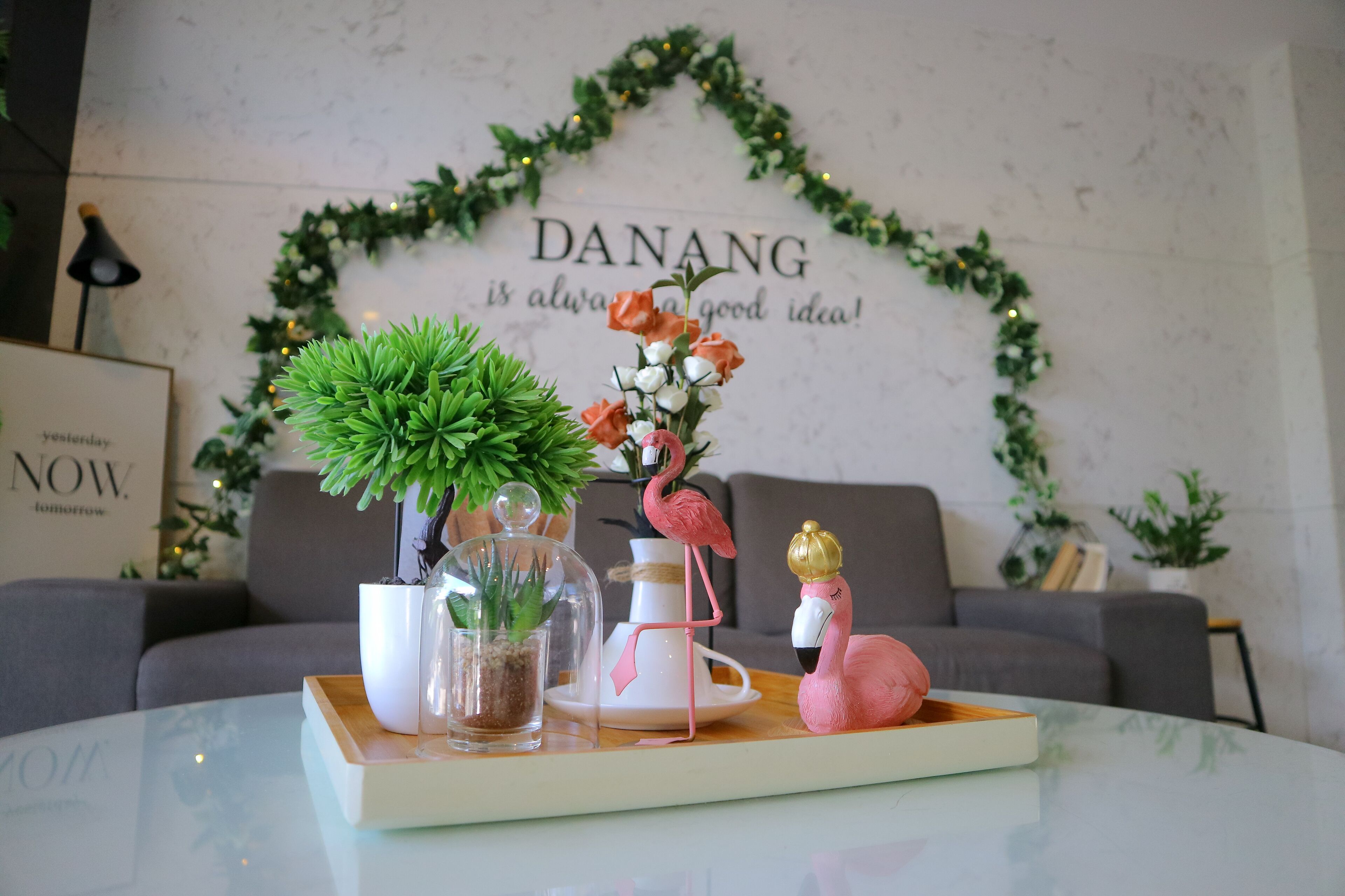 A table with a glass top displaying a potted plant, a glass dome, a white vase, and a pink flamingo figurine.