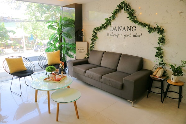 Una sala de estar moderna con sofá, mesa de centro de vidrio y sillas, una pared decorativa con follaje y un letrero que dice "DANG is always a good idea!"