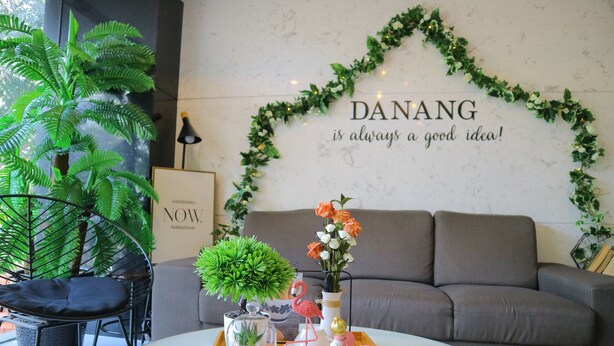 A cozy living area with a grey sofa, a round coffee table, and decorative items including a flamingo figurine, a vase with flowers, and a potted plant. A wall with a heart-shaped garland and a motivational quote is visible. - Robin Hotel Danang (Da Nang)