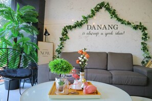 A cozy living area with a grey sofa, a round coffee table, and decorative items including a flamingo figurine, a vase with flowers, and a potted plant. A wall with a heart-shaped garland and a motivational quote is visible. - Robin Hotel Danang (Da Nang)