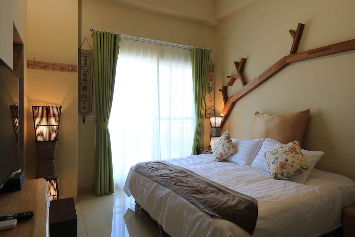 Una cama bien hecha con almohadas estampadas, un televisor sobre una mesa de madera y una lámpara fijada a la pared.