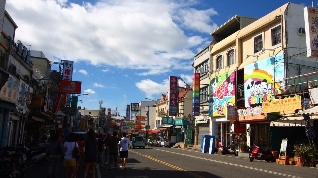 Une rue animée, bordée de boutiques et de panneaux de toutes sortes, dont une devanture colorée ornée d’un motif en arc-en-ciel.