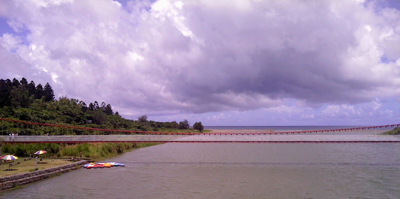 A riverside view with a bridge, trees, and a cloudy sky.