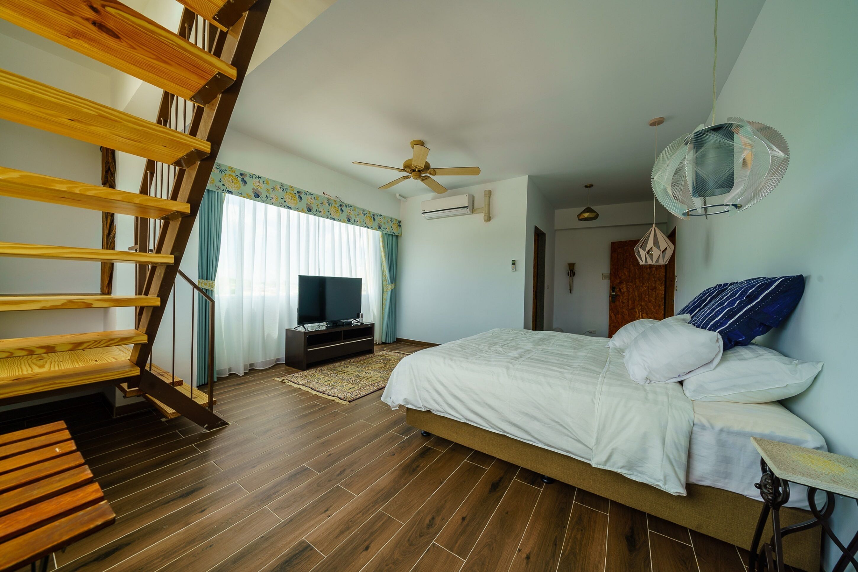 A bedroom with a wooden staircase, a bed with white bedding, a ceiling fan, and a TV on a stand.