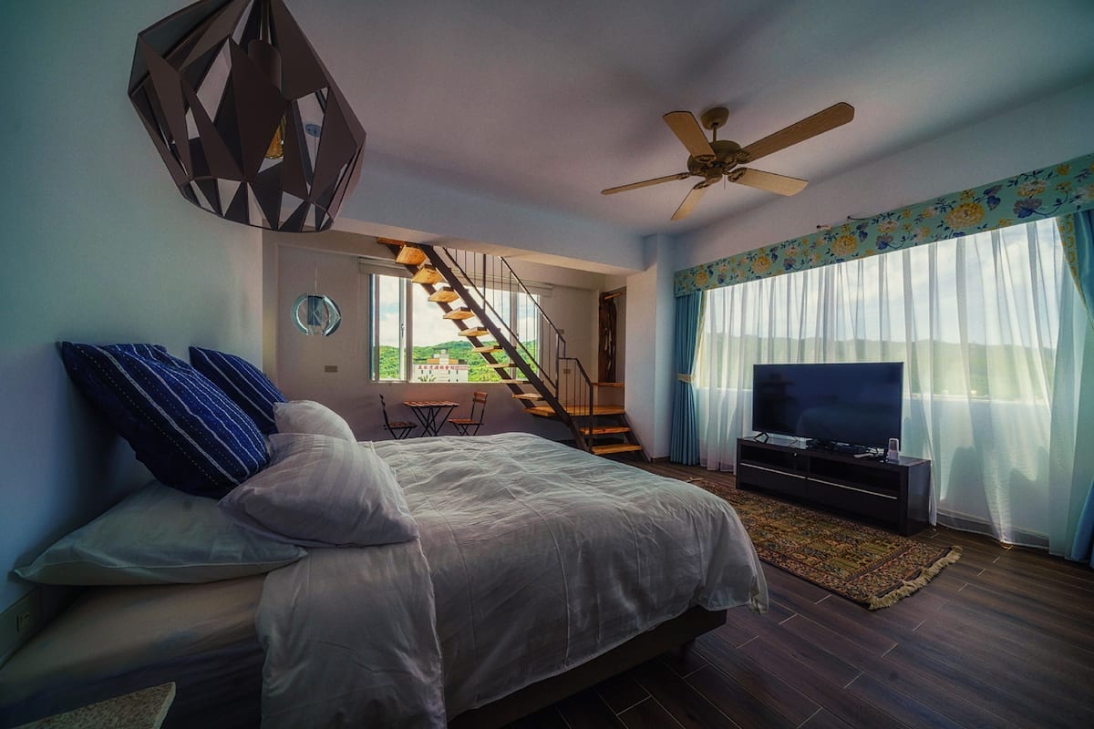 Un dormitorio con cama, televisión, ventilador de techo, escalera y vistas al exterior.