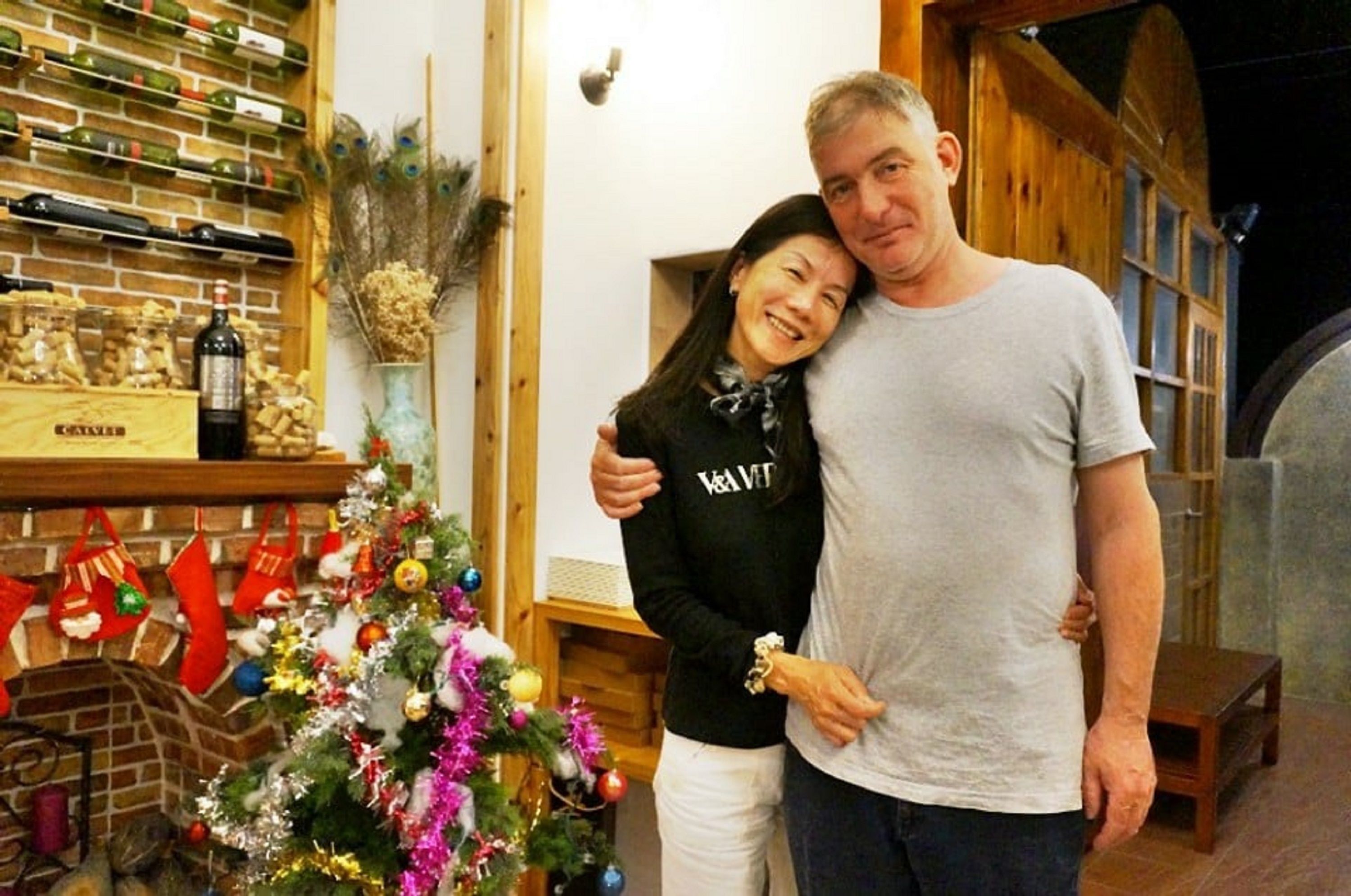 Um homem e uma mulher estão em um quarto com uma árvore de Natal, prateleiras de madeira com garrafas e uma lareira de tijolos.