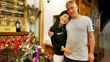 Um homem e uma mulher estão em um quarto com uma árvore de Natal, prateleiras de madeira com garrafas e uma lareira de tijolos.