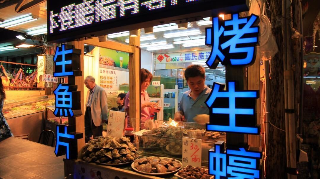 A seafood stall with a neon sign, displaying various seafood items for sale.
