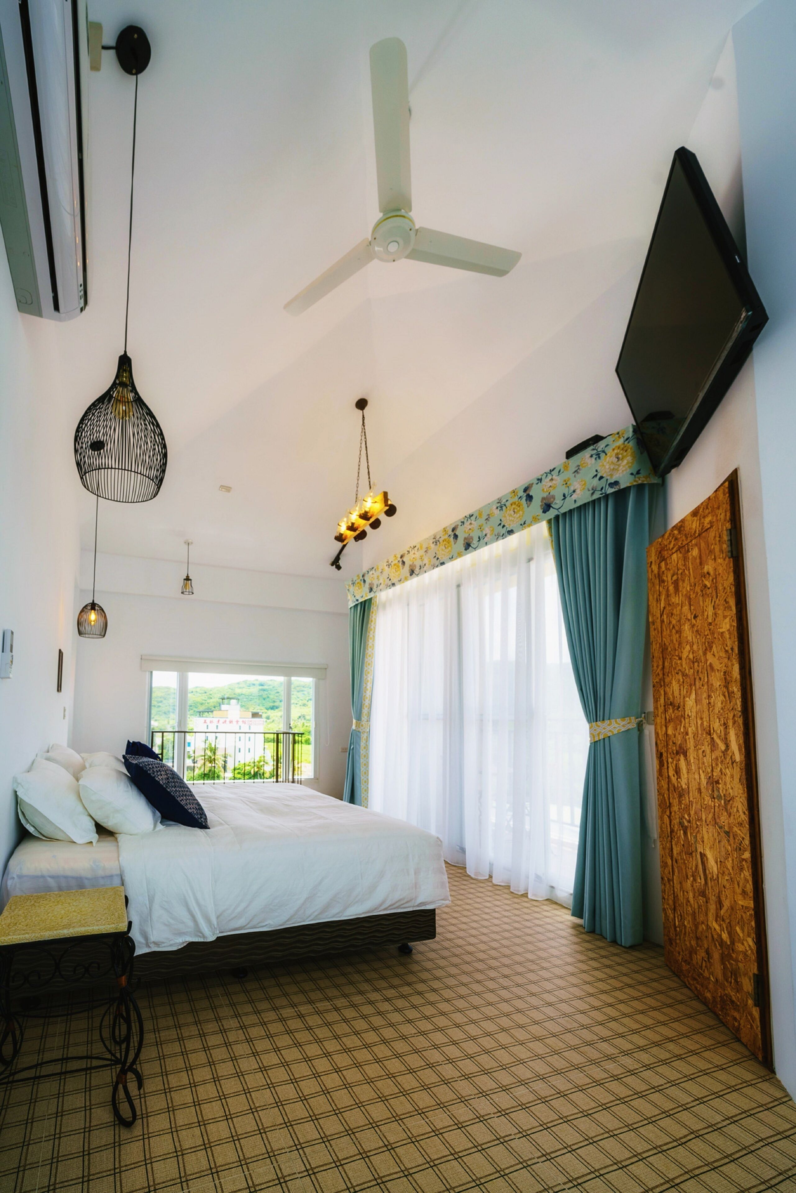 Un dormitorio con cama, ventilador de techo, televisor montado en la pared y ventana con cortinas.
