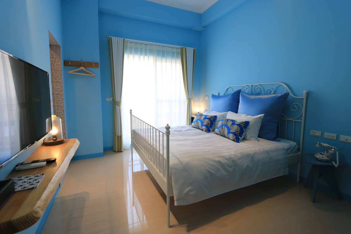 Un dormitorio con cama, televisor, mesita de noche y ventana con cortinas.