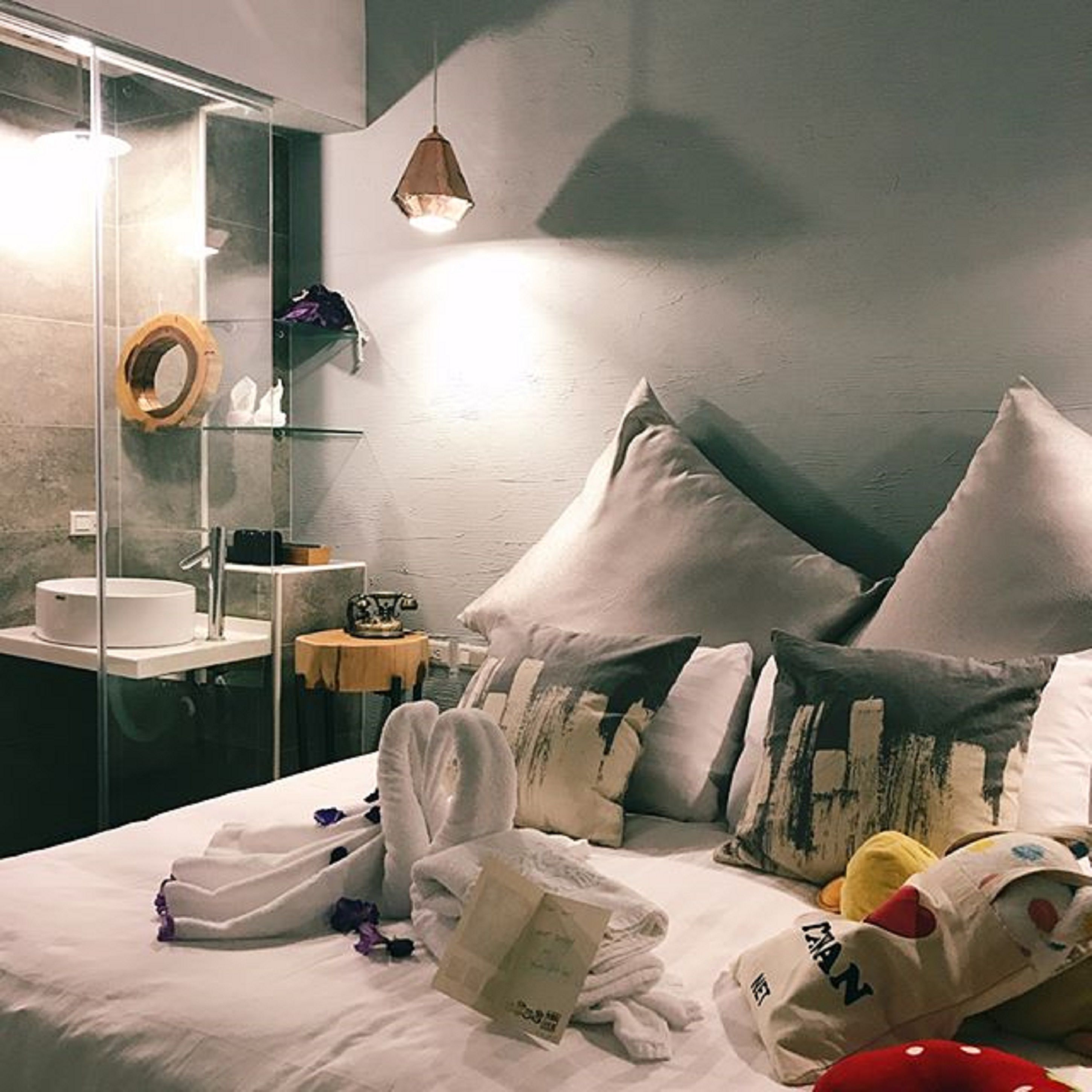 Habitación de hotel con cama, almohadas, cabina de ducha de vidrio, lavamanos y una pequeña mesa con lámpara.