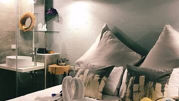 Habitación de hotel con cama, almohadas, cabina de ducha de vidrio, lavamanos y una pequeña mesa con lámpara.