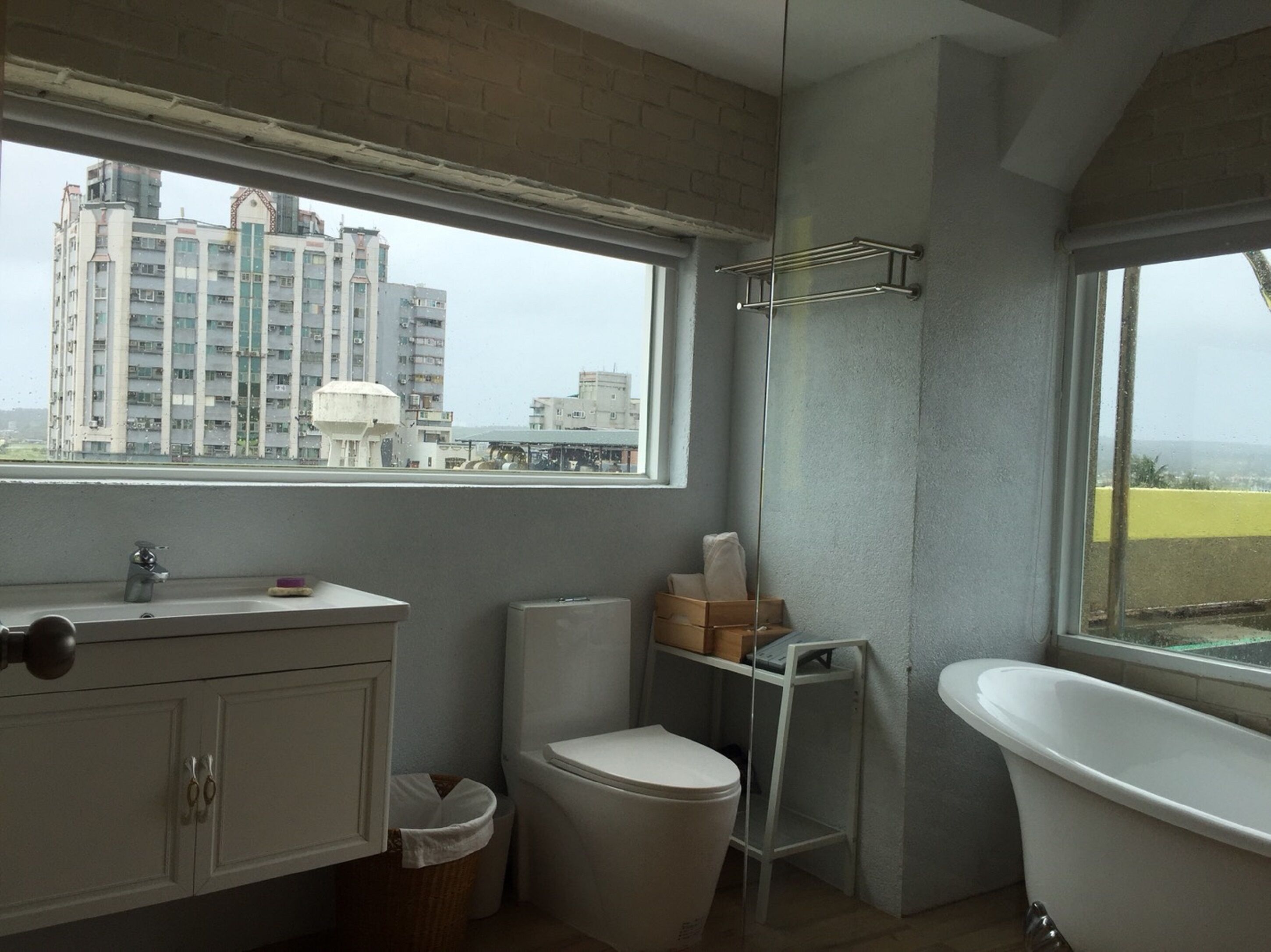 Un baño con inodoro, lavamanos y bañera. Se ve una ventana con vistas a edificios.