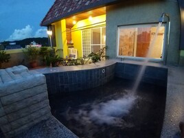 Un área de hidromasaje con piso de baldosas, terraza de madera y zona de estar techada con sillas.