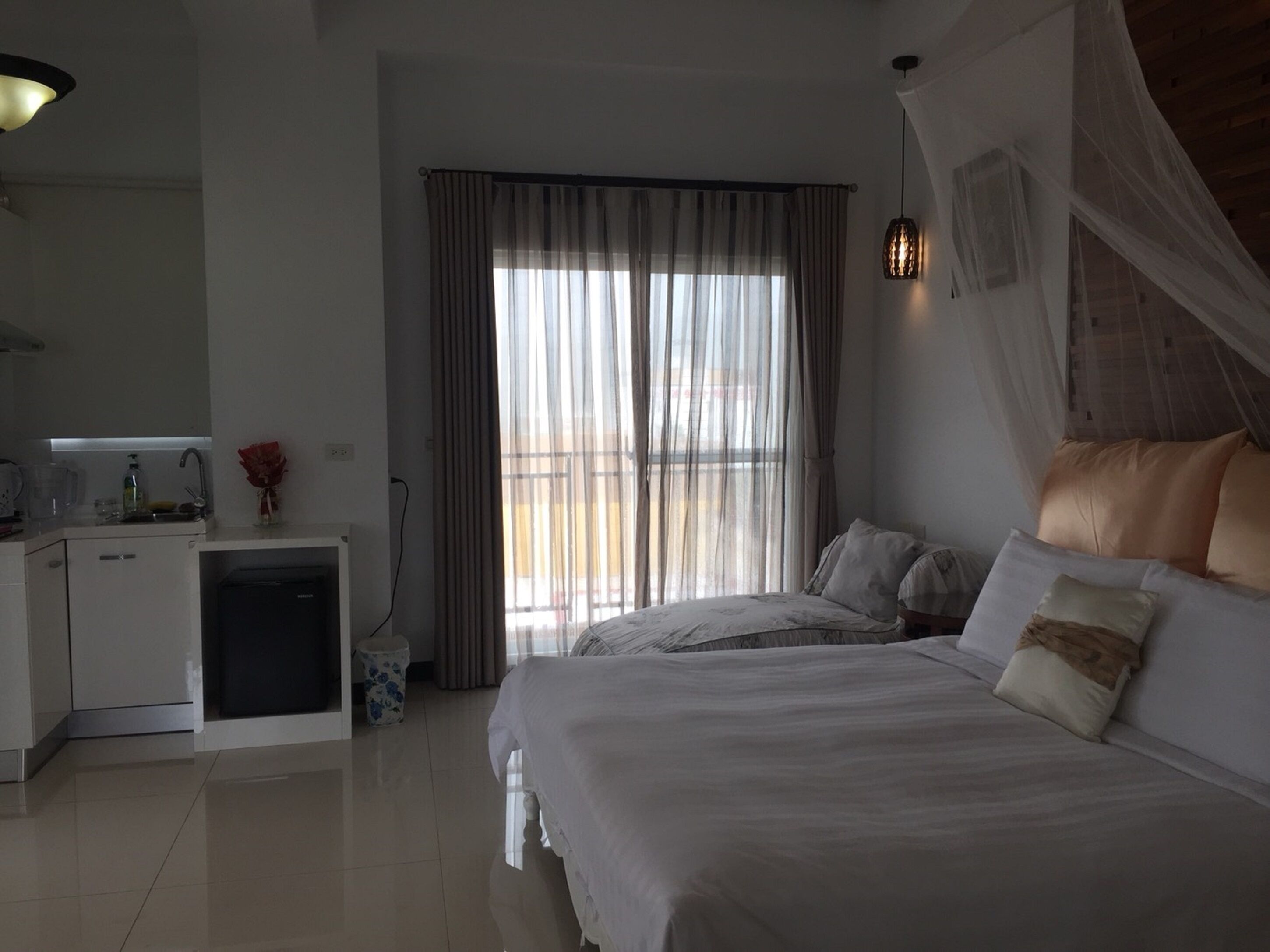 A hotel room with a bed, a bedside table, a small desk with a chair, a window with curtains, and a hanging light fixture.