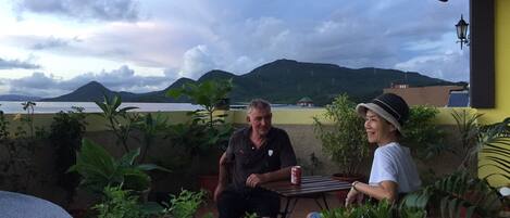 Two people seated at an outdoor table with a view of the sea and mountains in the background.