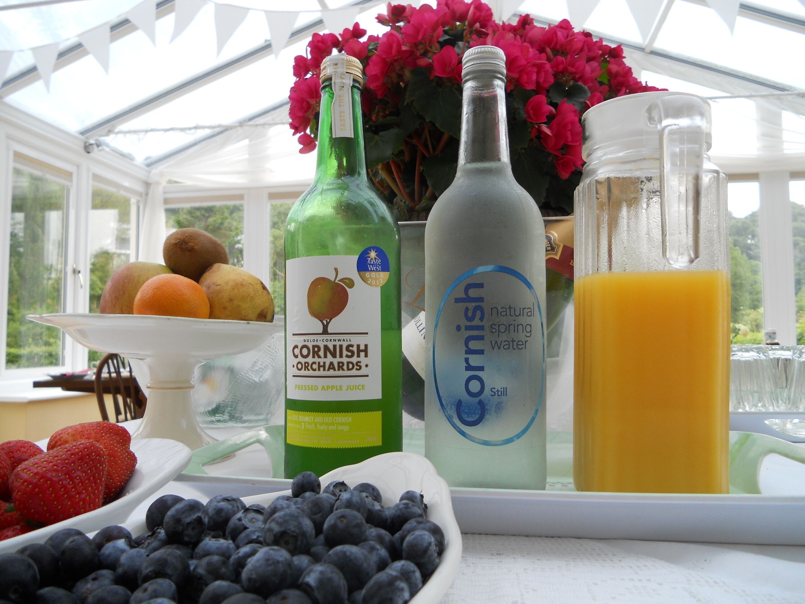 A table with a bottle of pressed apple juice, a bottle of natural spring water, a glass of orange juice, and bowls of strawberries and blueberries.