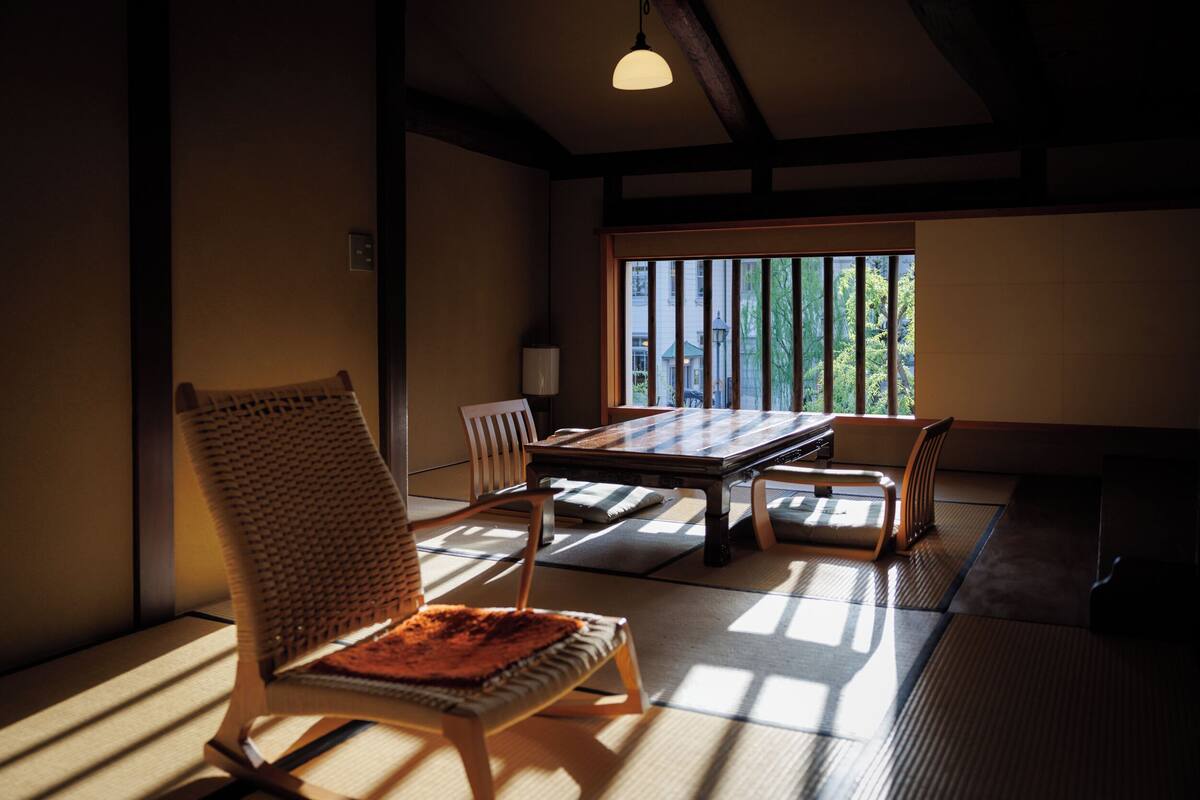 Ein traditioneller japanischer Raum mit Tatami-Boden, einem niedrigen Tisch, Holzstühlen und Blick durch einen Shoji-Bildschirm.