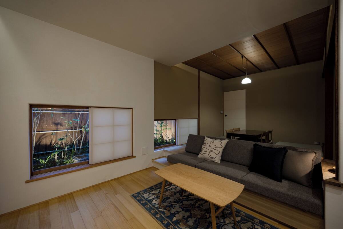 Ein Wohnzimmer mit einer Couch, einem Holztisch, einem gemusterten Teppich und einer Glastür mit Blick ins Grüne.