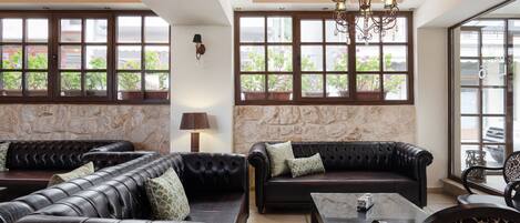 A modern lounge area with leather sofas, a glass coffee table, and a chandelier. Large windows with potted plants are visible.