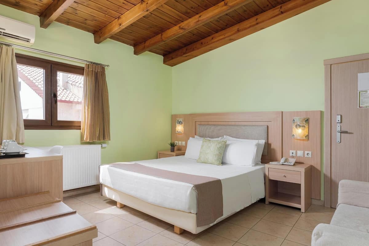 Ein Hotelzimmer mit Holzboden, einem Bett mit weißer Bettwäsche, einem Nachttisch mit Lampe, einem Stuhl und einem Fenster mit Vorhängen.