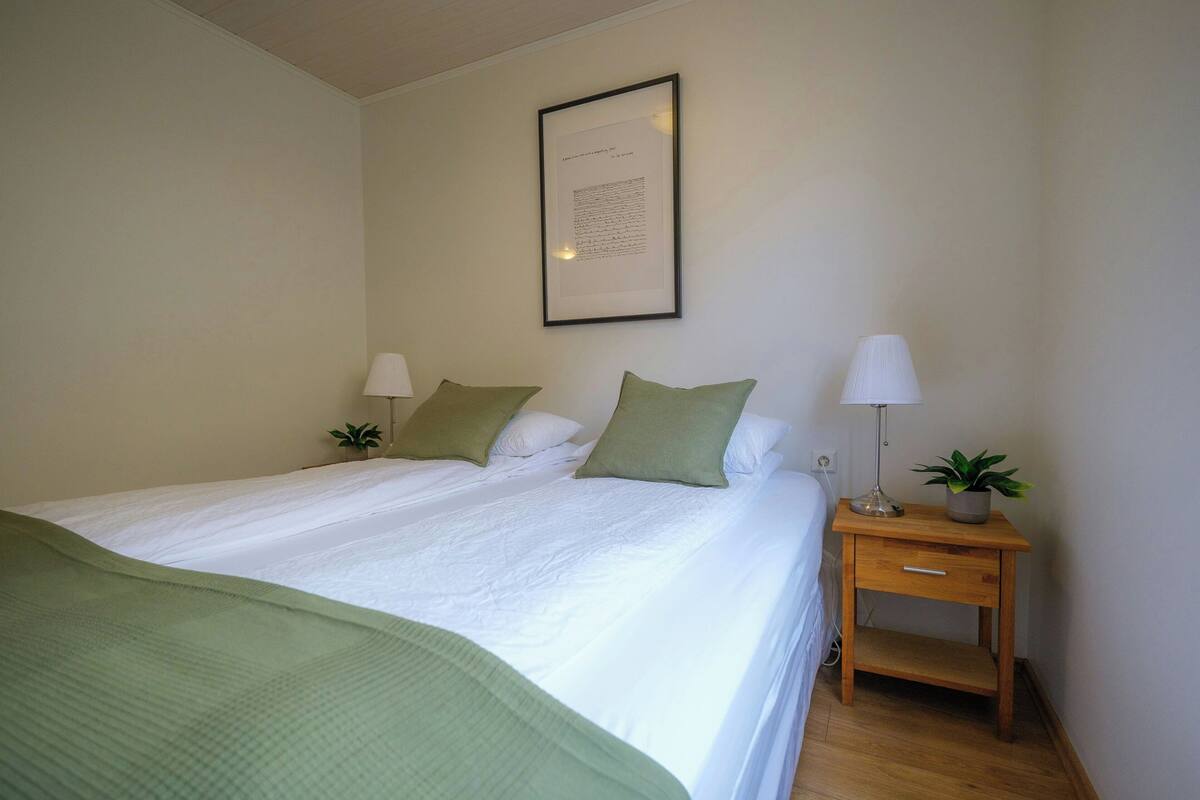 A neatly made bed with pillows, a bedside table with a lamp, and a framed document on the wall.