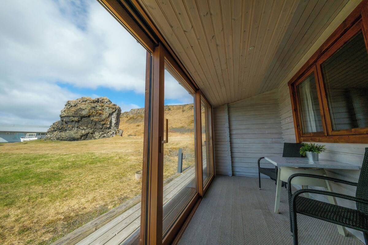 Ein Zimmer mit großen Fenstern, einer Holzbalkendecke, einem Tisch mit Pflanze und Blick auf eine felsige Landzunge.