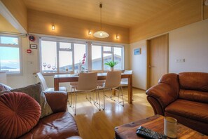 Uma sala de estar com um sofá de couro marrom, uma mesa de centro de madeira, uma mesa de jantar com cadeiras e uma janela grande com vista.