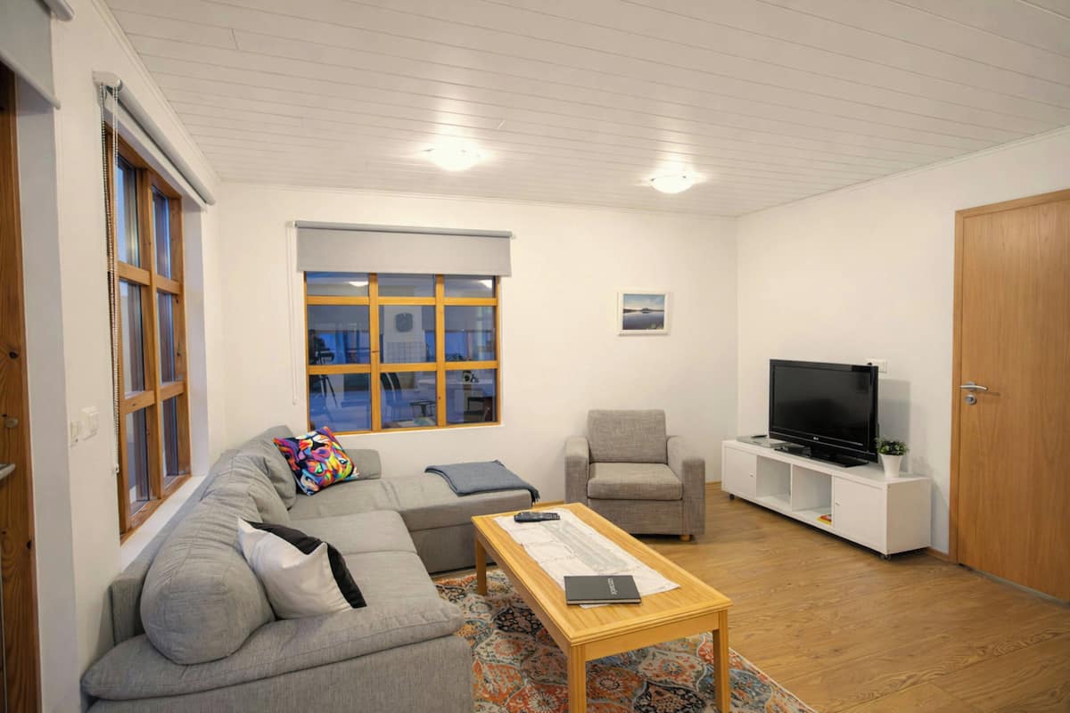 A living room with a gray sofa, a small armchair, a coffee table, a TV on a white stand, and a window with blinds.