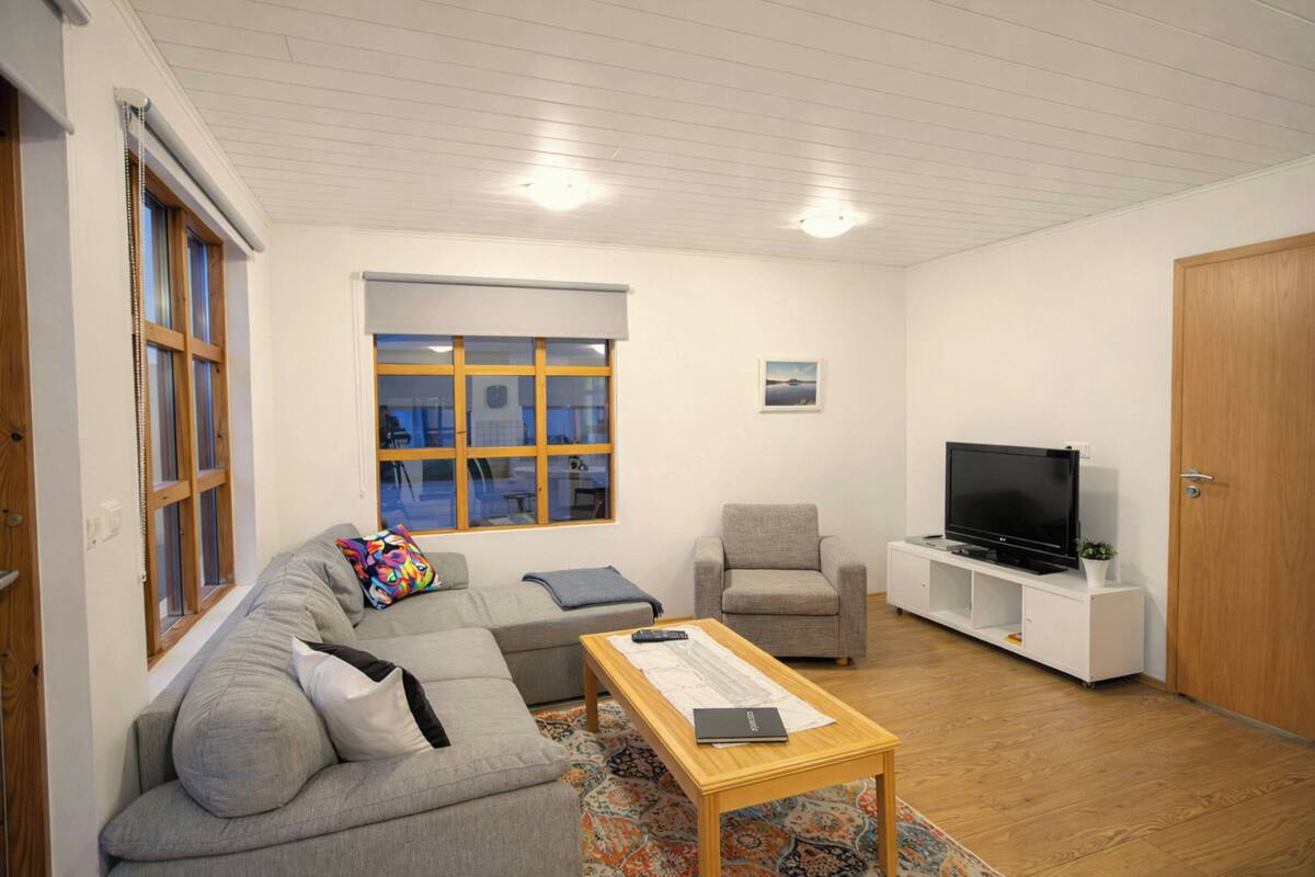 Ein Wohnzimmer mit grauem Sofa, einem kleinen Sessel, einem Couchtisch, einem Fernseher auf einem weißen Schrank und einem Fenster mit Jalousien.