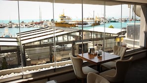 A dining area with a view of a harbor, featuring a table and chairs, and a large window overlooking the water. - Sinop Mola Otel (Sinop)