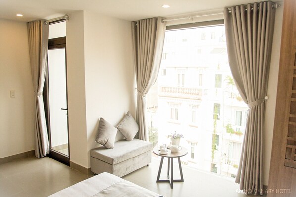 A hotel room with a sofa, a small table with a teapot, and a view of buildings through large windows. - An Hoi Canary Hotel (Da Nang)