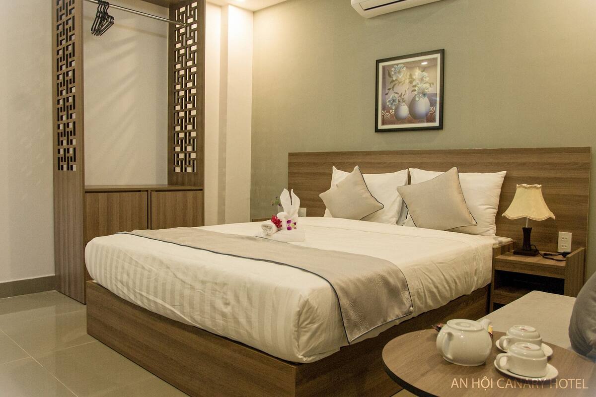 A hotel room with a wooden bed, a nightstand with a lamp, and a small table with a teapot and cups.