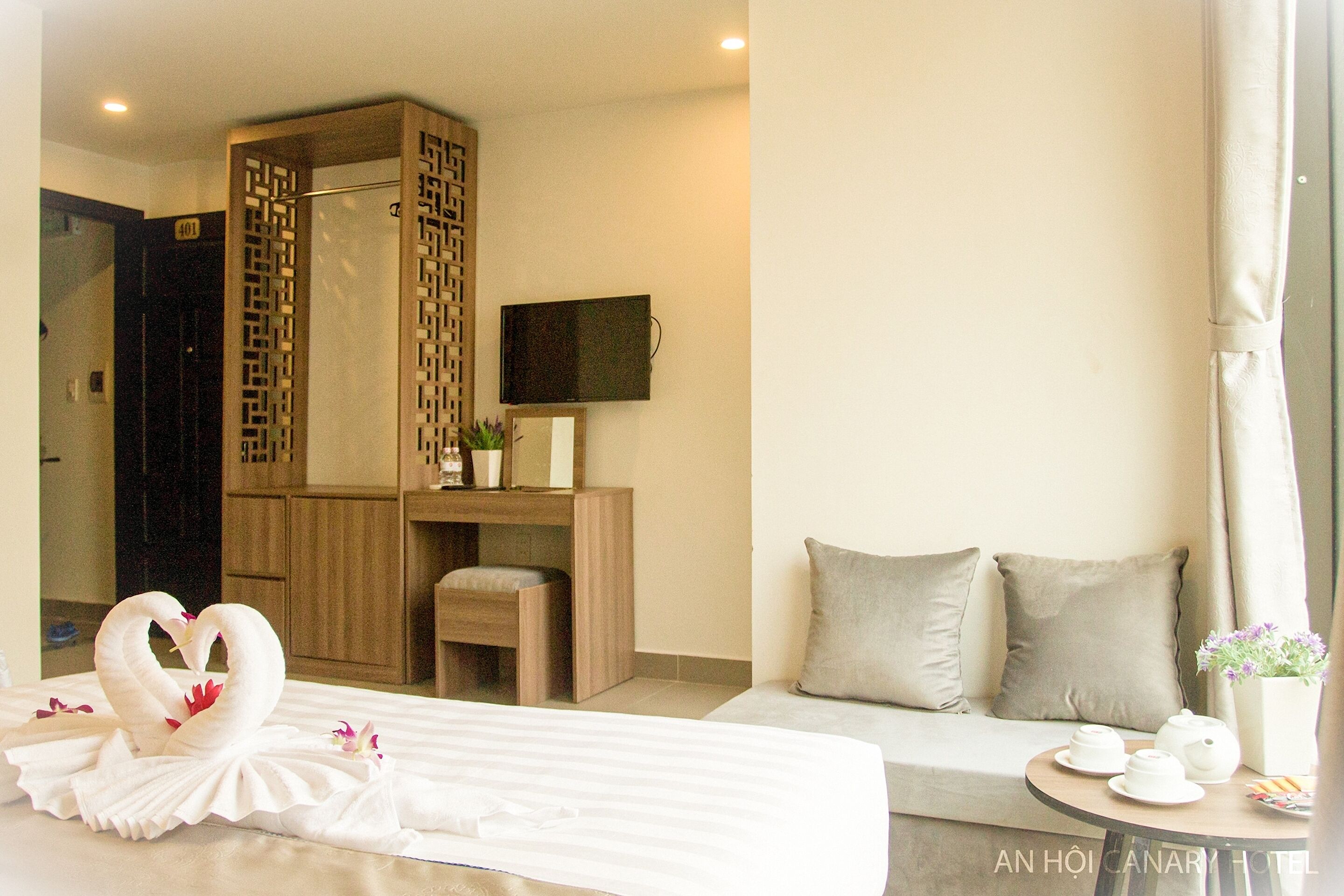 A hotel room with a bed, a sofa, a desk with a chair, a TV, and a small table with cups and a flower vase.