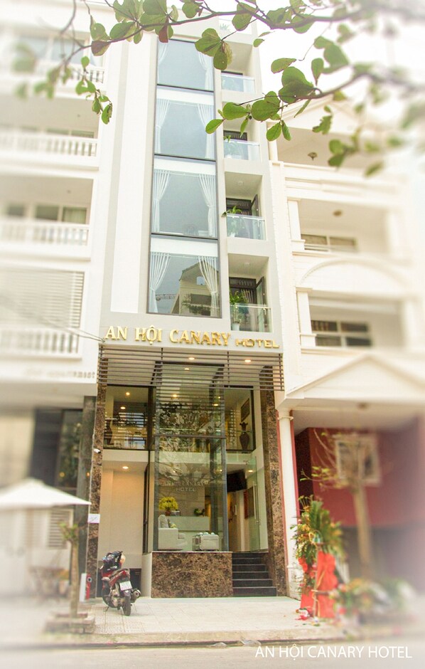 Vue de la façade de l’hôtel An Hoi Canary, avec une vue dégagée sur l’entrée, une moto garée à l’extérieur et un pot de fleurs sur le côté droit.
