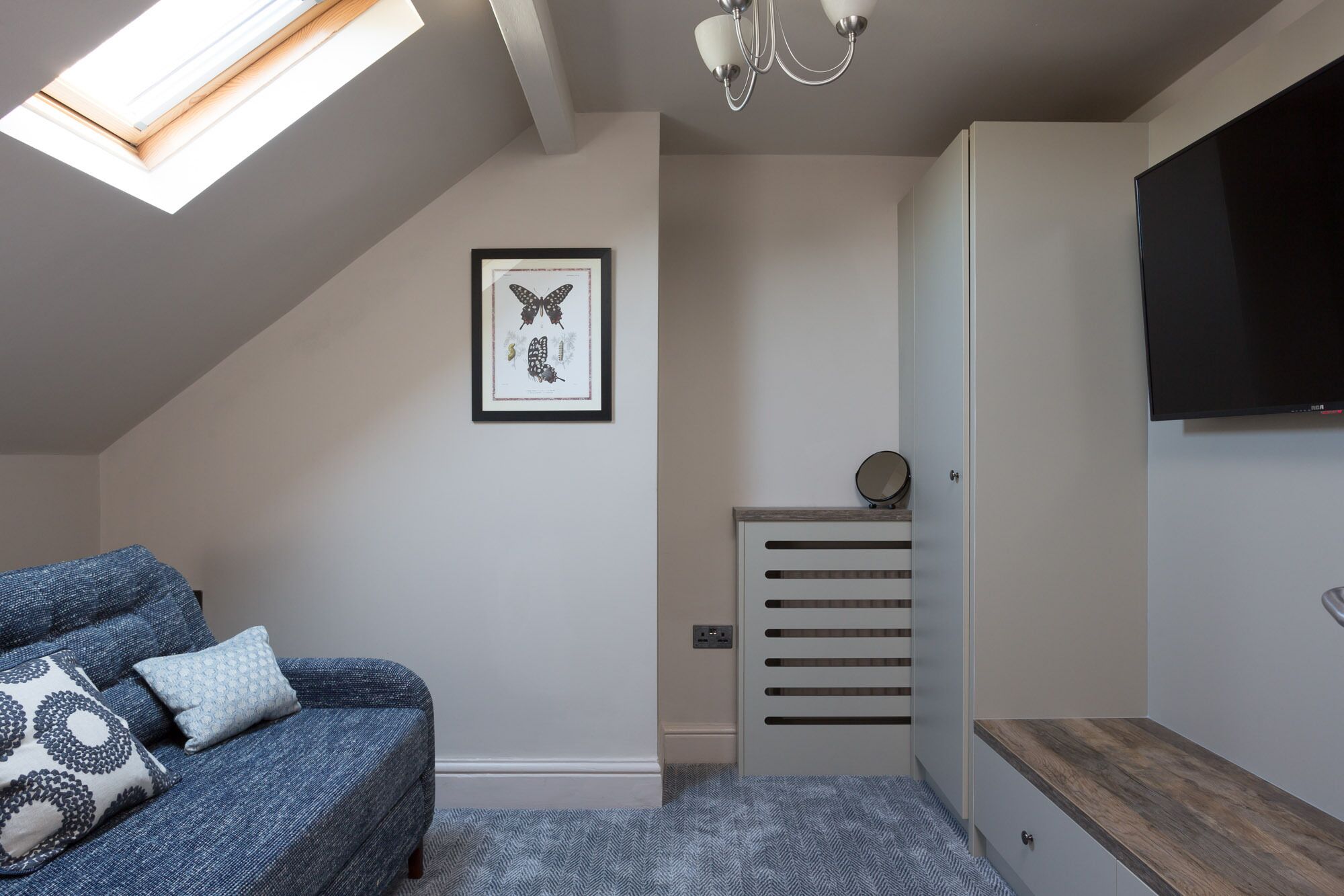 A modern bedroom with a sloped ceiling, a skylight, a framed butterfly artwork, a bedside table with a mirror, and a TV mounted on the wall.