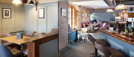 A restaurant with a bar area, wooden tables, and chairs. There are framed pictures on the walls and a stone wall in the background.