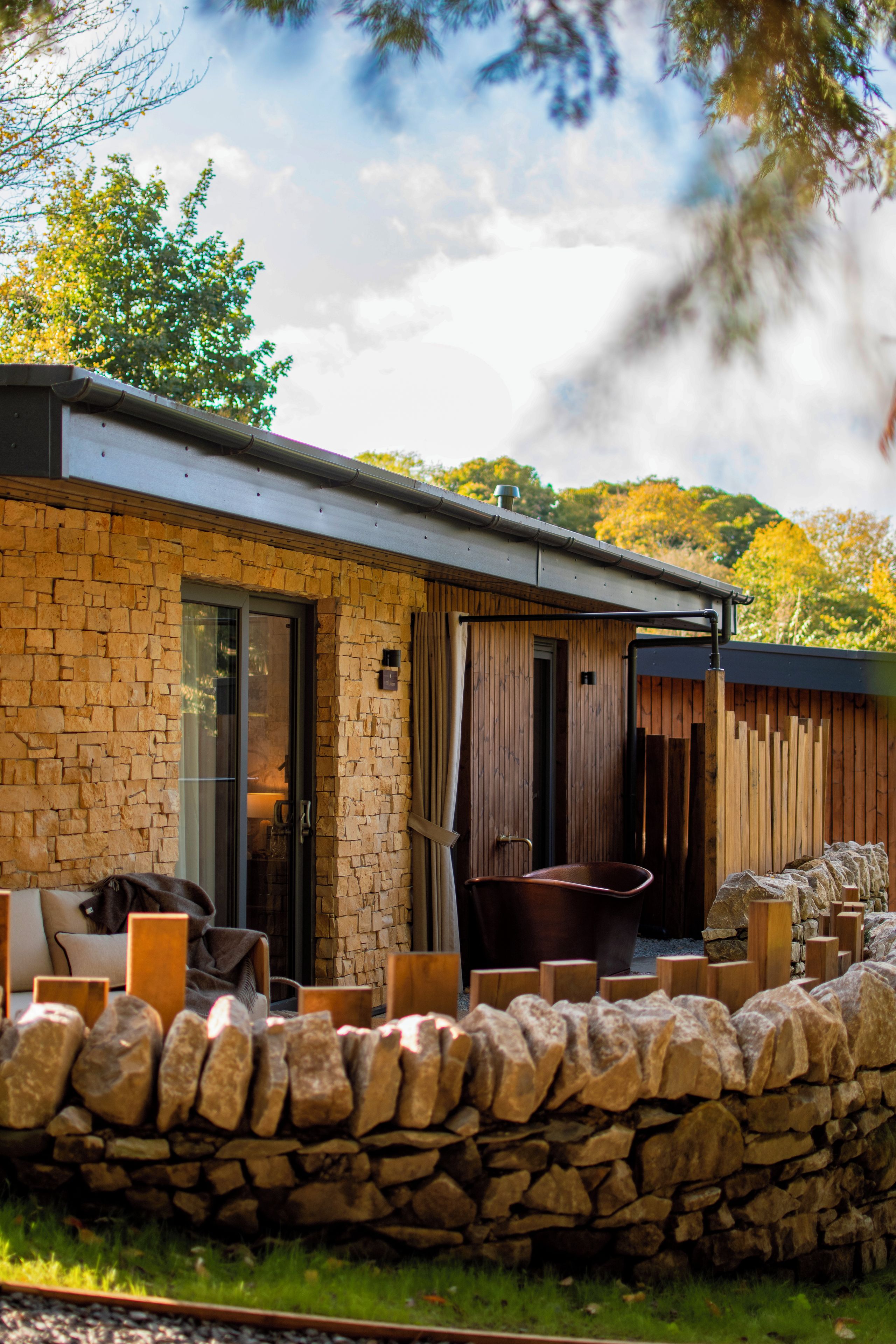 A modern building with stone walls, a glass door entrance, and a seating area with a stone wall and wooden furniture.