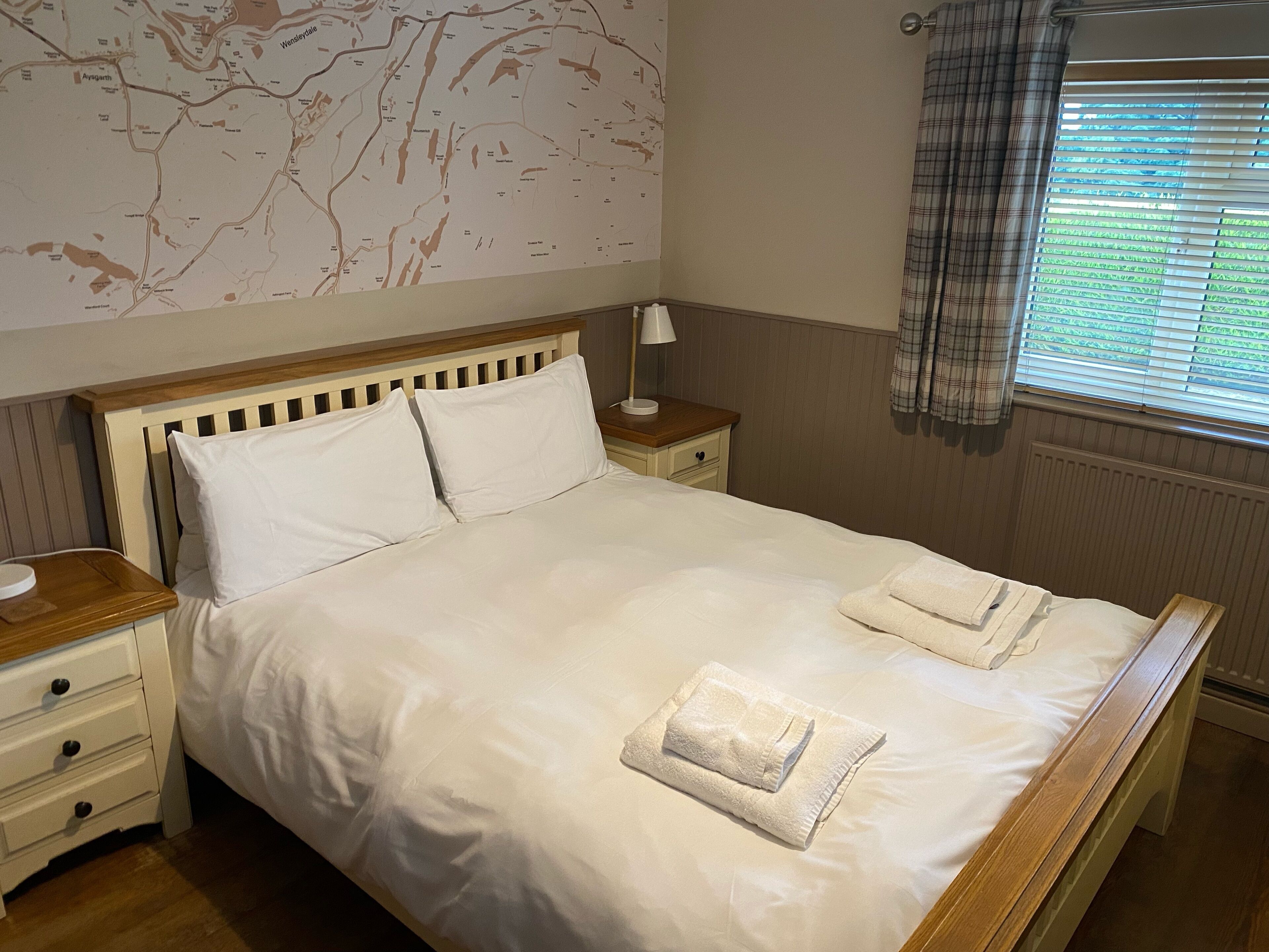 A neatly made bed with white linens and two folded towels, a wooden headboard, a nightstand with a lamp, and a map on the wall.