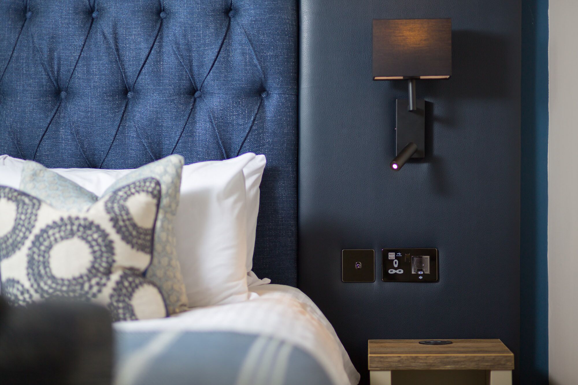 A bedroom with a tufted headboard, a bedside table with a lamp, and a wall-mounted control panel.