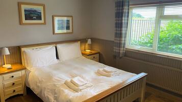 A neatly made bed with white linens, two bedside tables with lamps, a window with blinds, and two framed pictures on the wall.