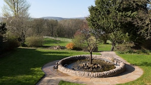 A circular stone fountain with a central tree, surrounded by a stone pathway, set in a grassy area with trees and a distant fence. - Aysgarth Falls Hotel (Leyburn)