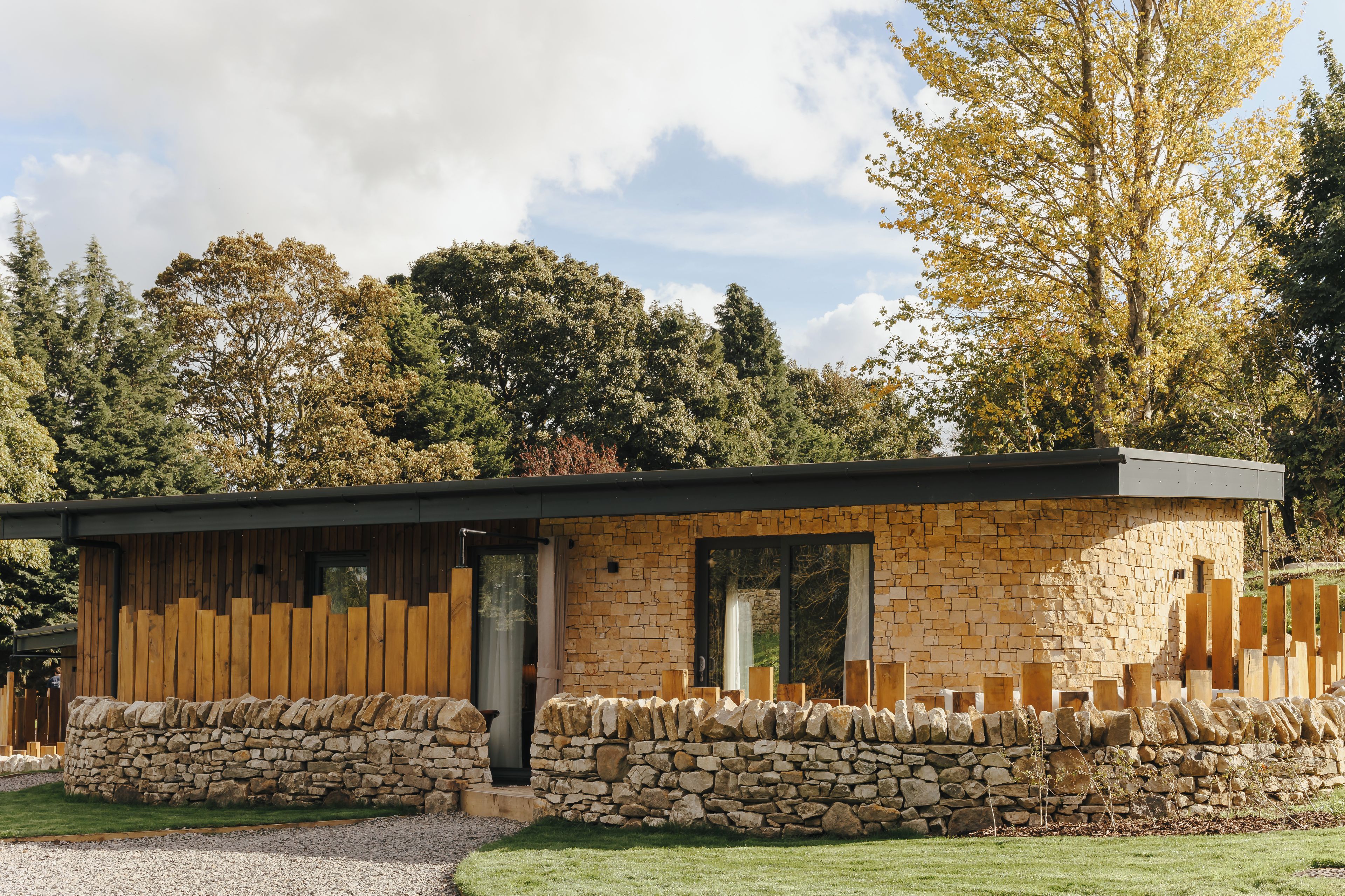 A modern stone building with a wooden deck and stone walls, surrounded by trees.