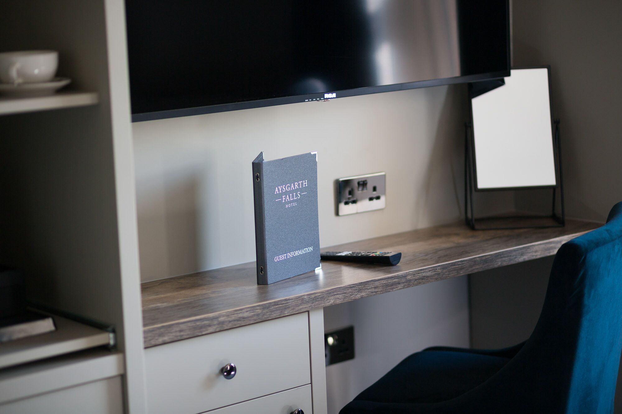 Habitación de hotel con escritorio, control remoto, un libro titulado 'Aysgarth Falls - Hotel Guest Information' y televisor.