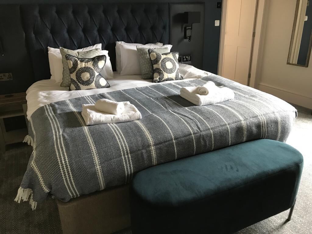 Una cama bien tendida con un edredón a rayas y dos almohadas, un cabecero oscuro y un banco color turquesa.