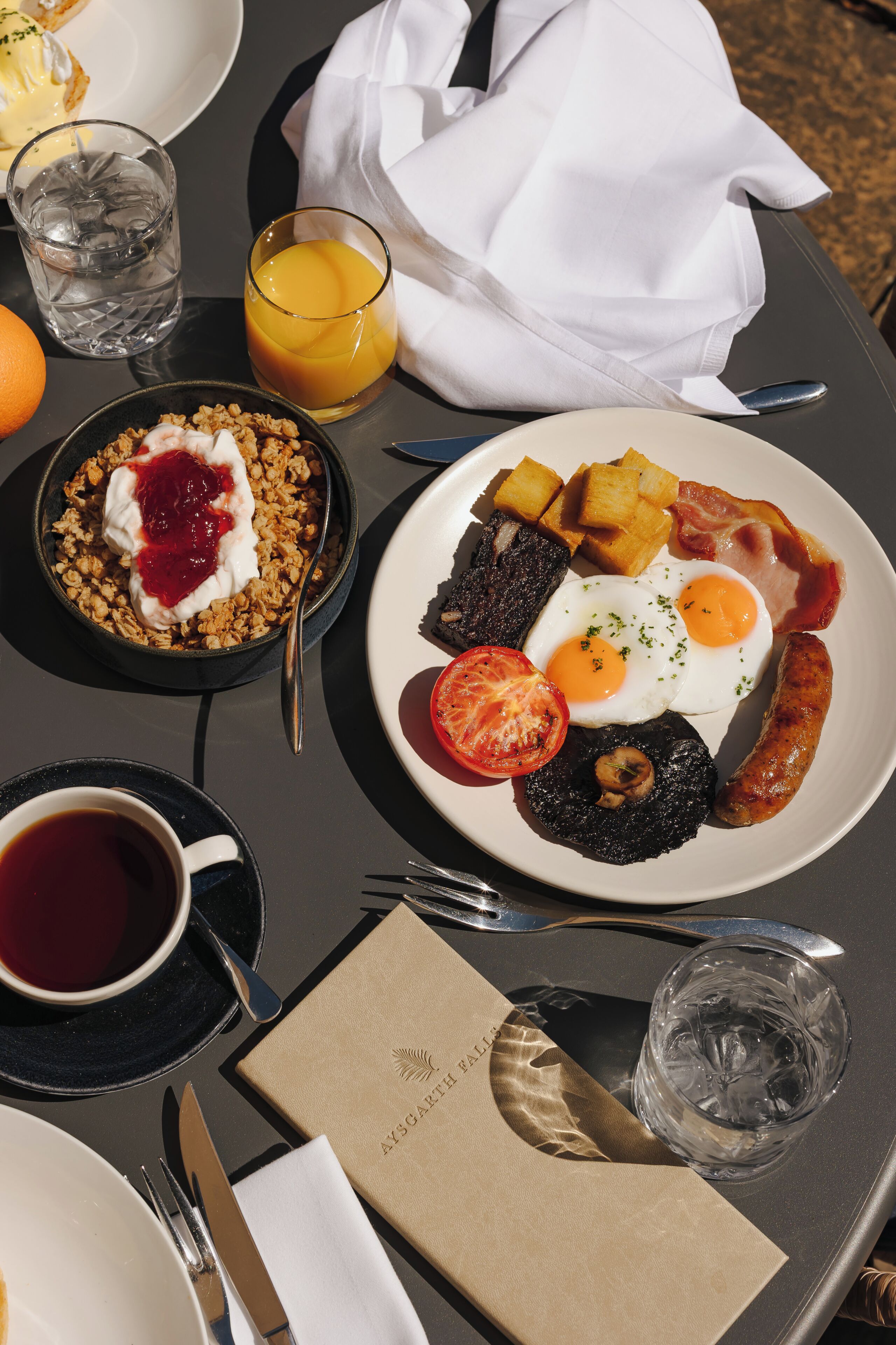 A breakfast setting with a plate of eggs, bacon, tomatoes, and toast, a bowl of cereal with yogurt, a glass of orange juice, and a cup of coffee.