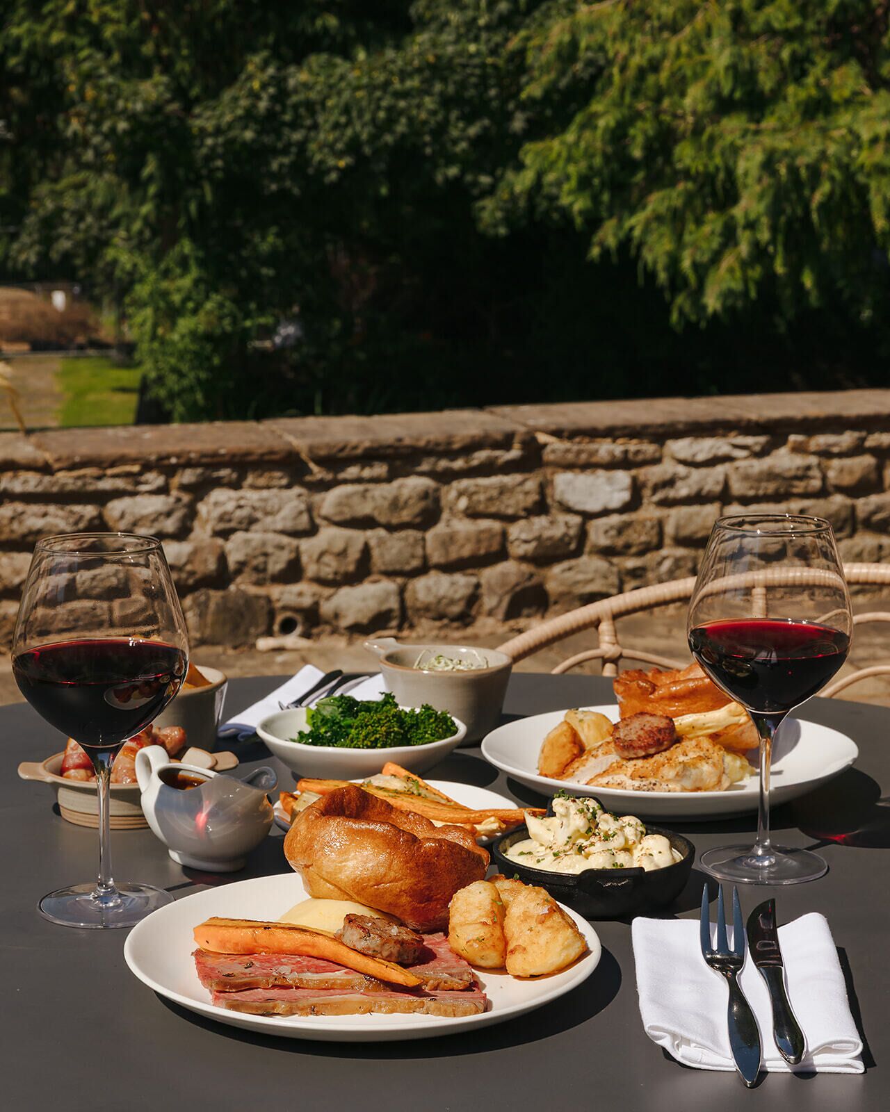 Una mesa servida con platos de comida, dos copas de vino tinto y una silla de mimbre en un jardín.